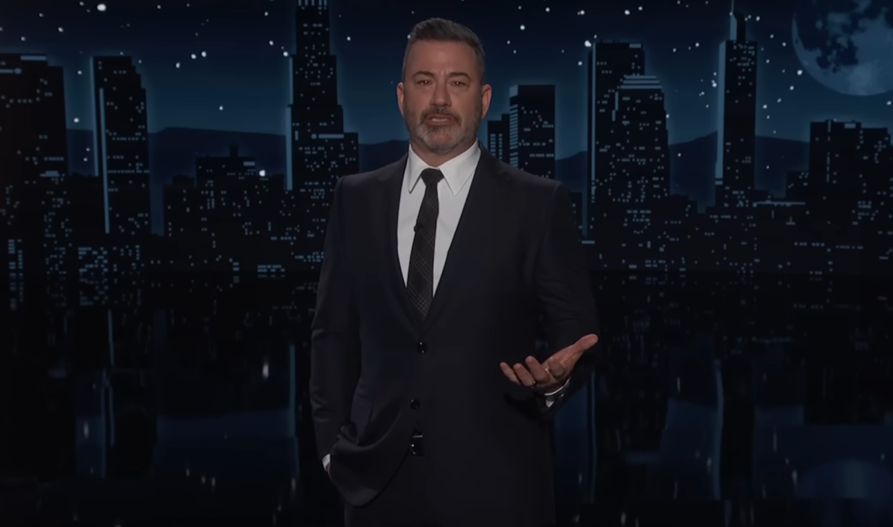 En mann i dress står på en scene med et bybilde i bakgrunnen, og gestikulerer med én hånd, sannsynligvis under en tale eller presentasjon