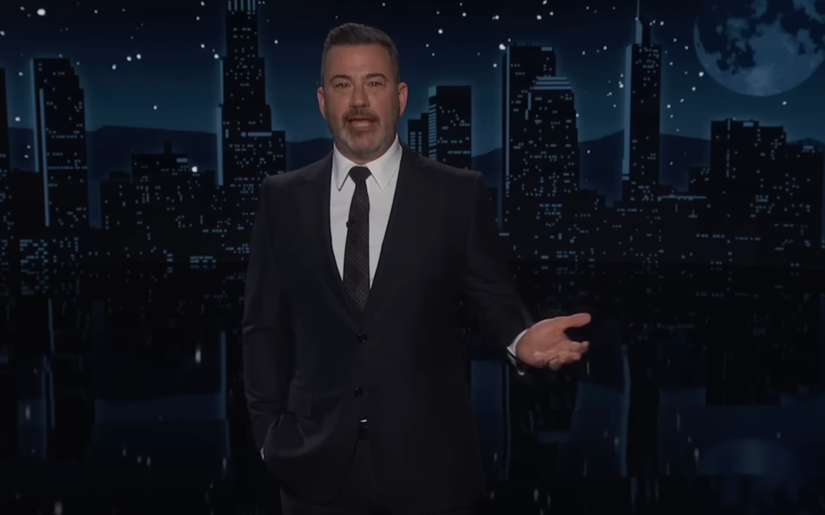 En mann i dress står på et talkshowsett med en bysilhuett i bakgrunnen