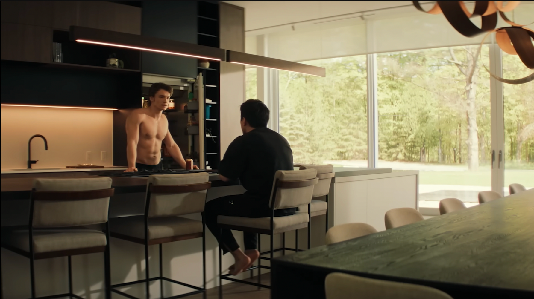 Two people in a modern kitchen; one is shirtless, standing at the counter, the other is seated on a stool, engaging in conversation