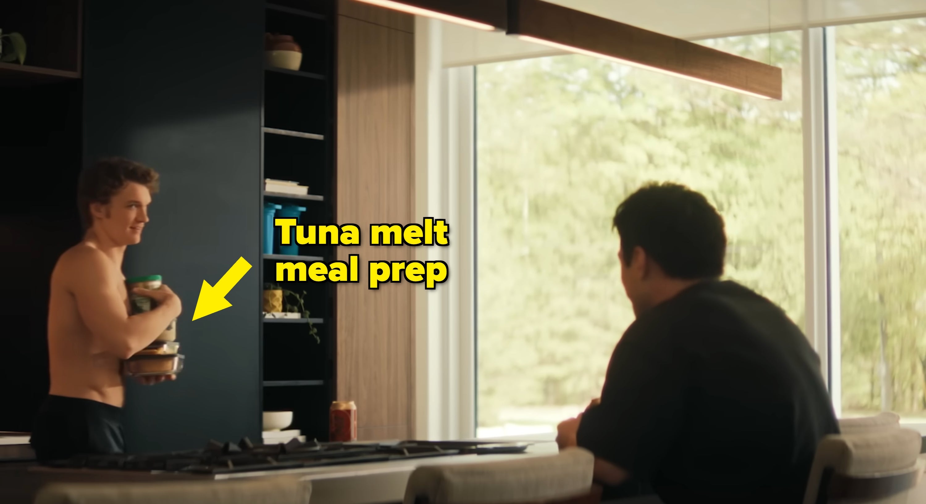 Two people in a modern kitchen. One is shirtless, holding jars. The other sits at a counter, facing away, engaging in a relaxed conversation
