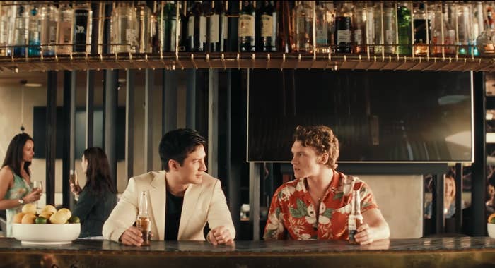 Two people sit at a bar with drinks, engaged in conversation. Others socialize in the background