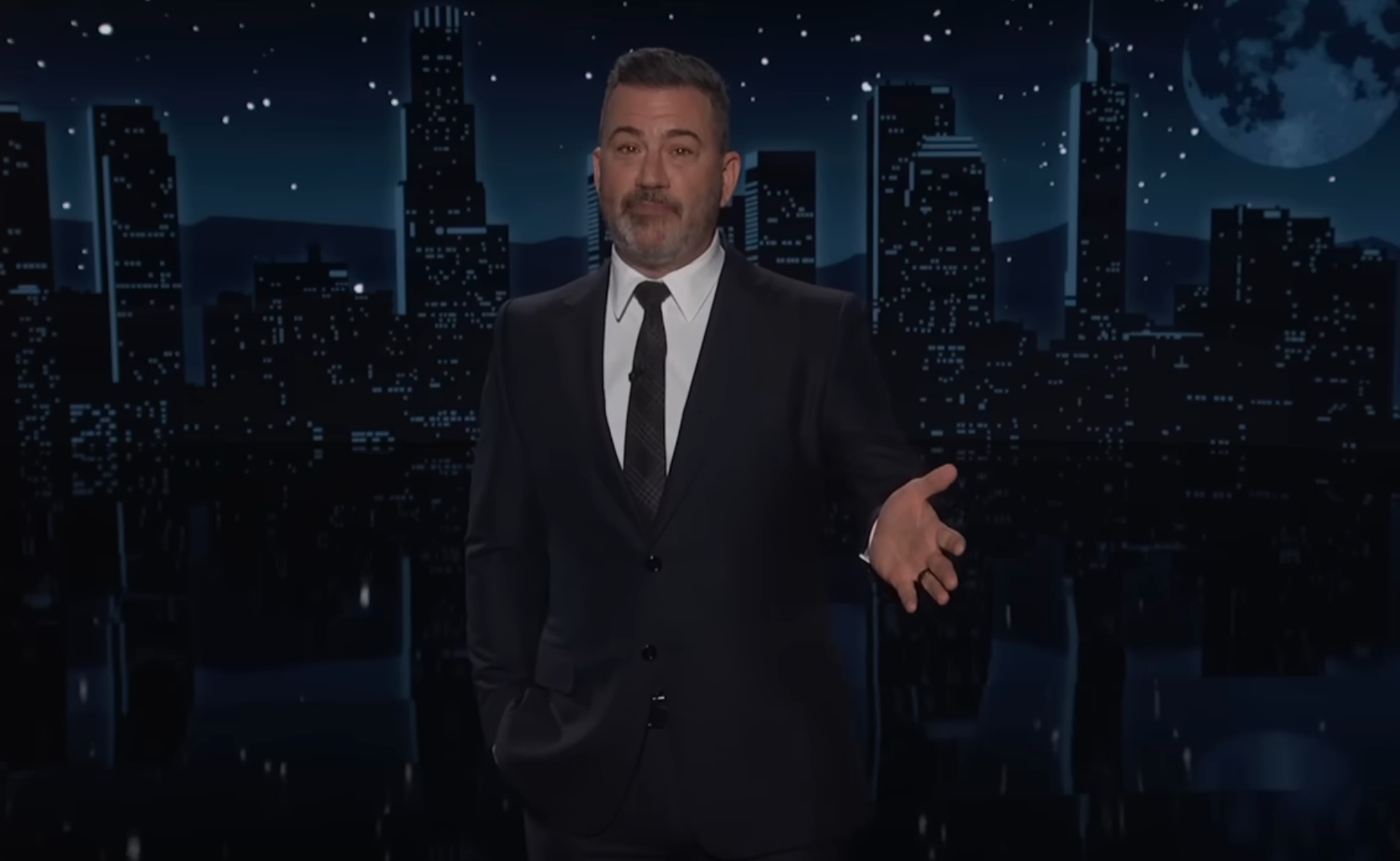 En mann i dress står foran en nattlig skyline i bakgrunnen, snakker eller presenterer