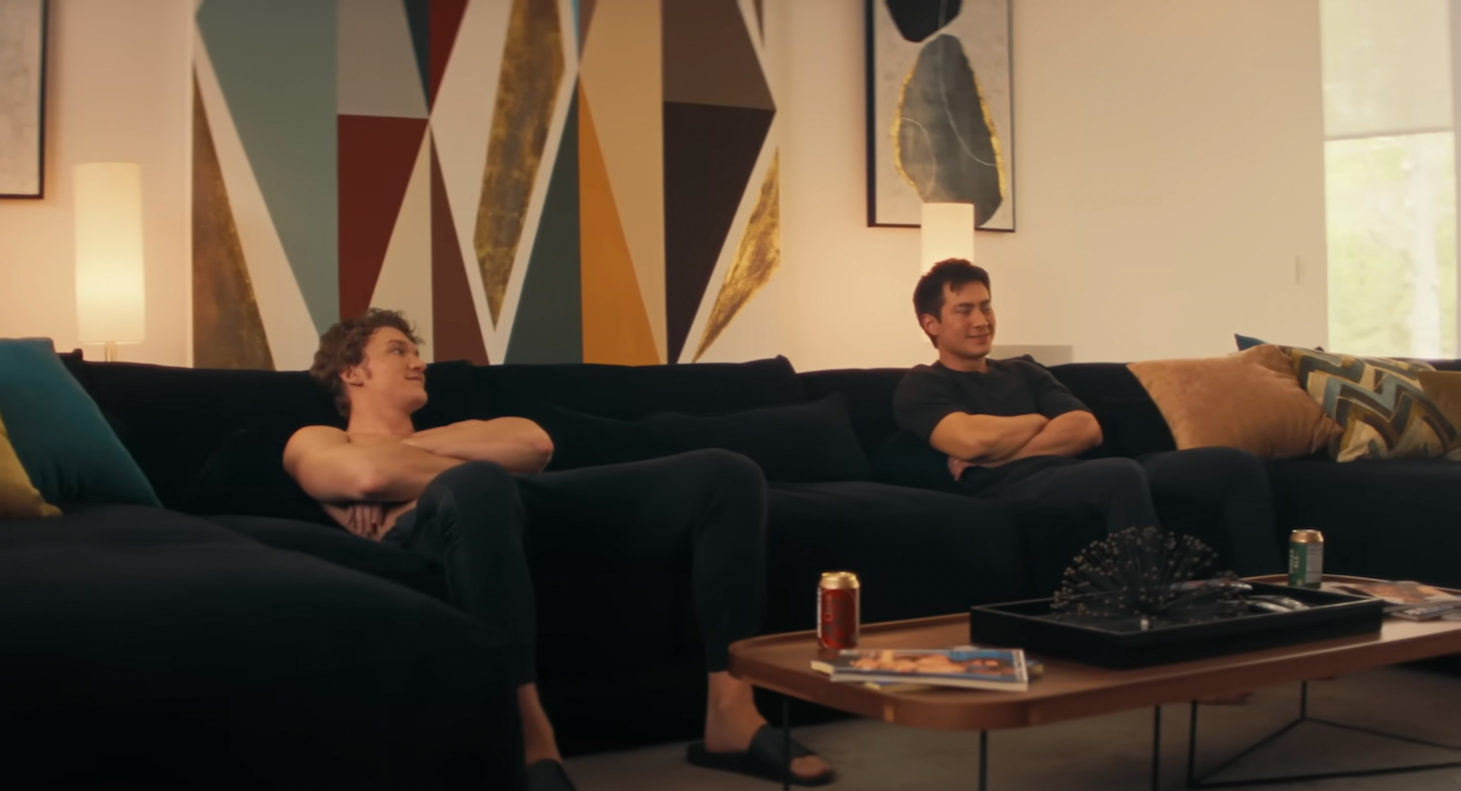 Two people are sitting on a black sofa in a modern living room, with geometric art on the wall behind them. There are drinks on the coffee table