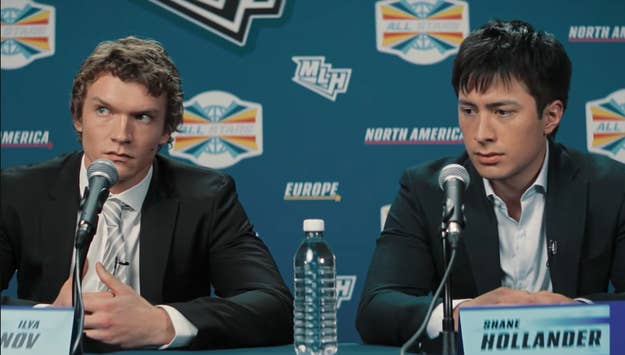 Two men in suits sit at a press conference table with microphones, appearing focused and attentive. A water bottle is on the table between them