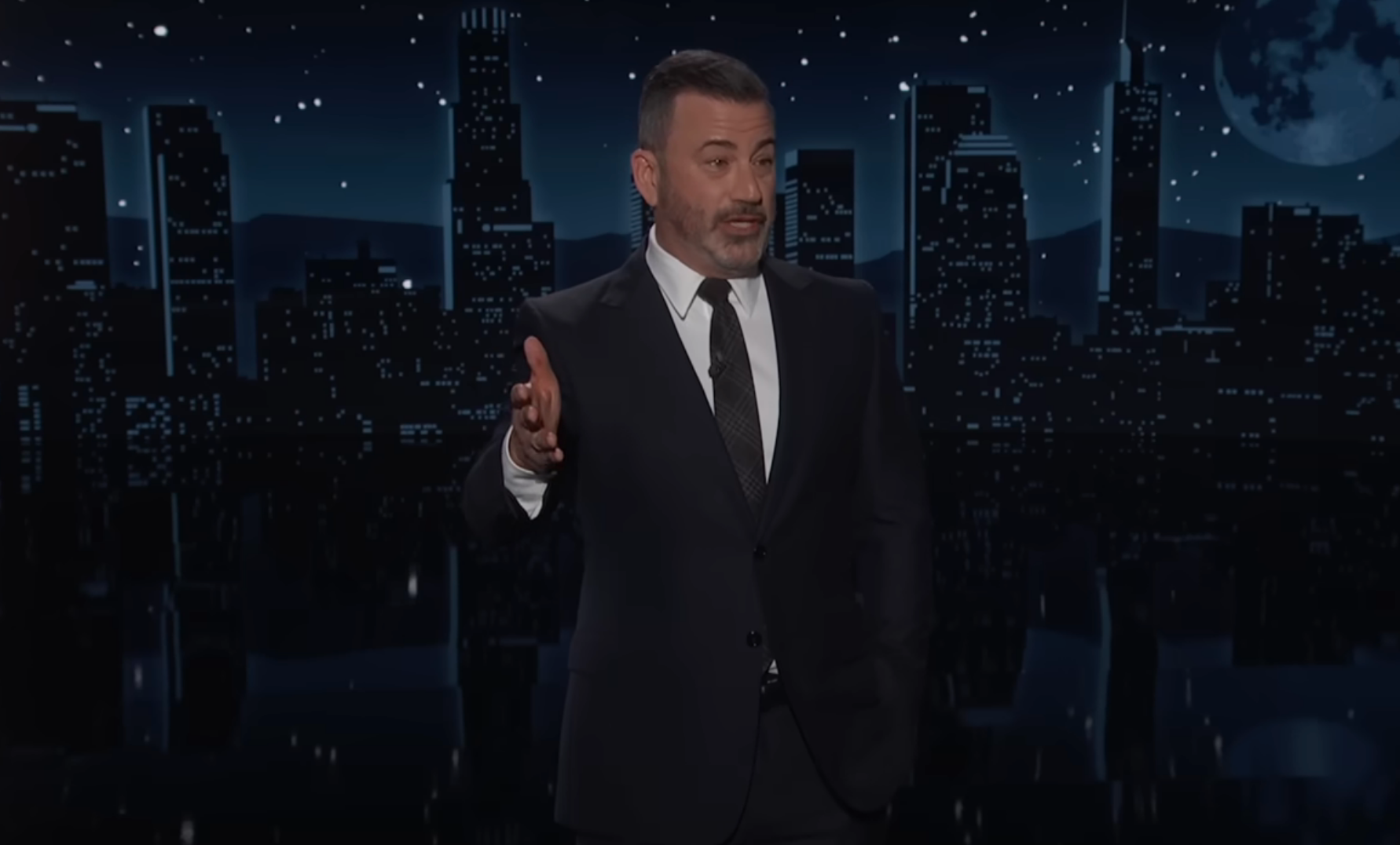 En mann i dress gestikulerer mens han snakker på et nattlig talkshowsett med en byhorisont i bakgrunnen