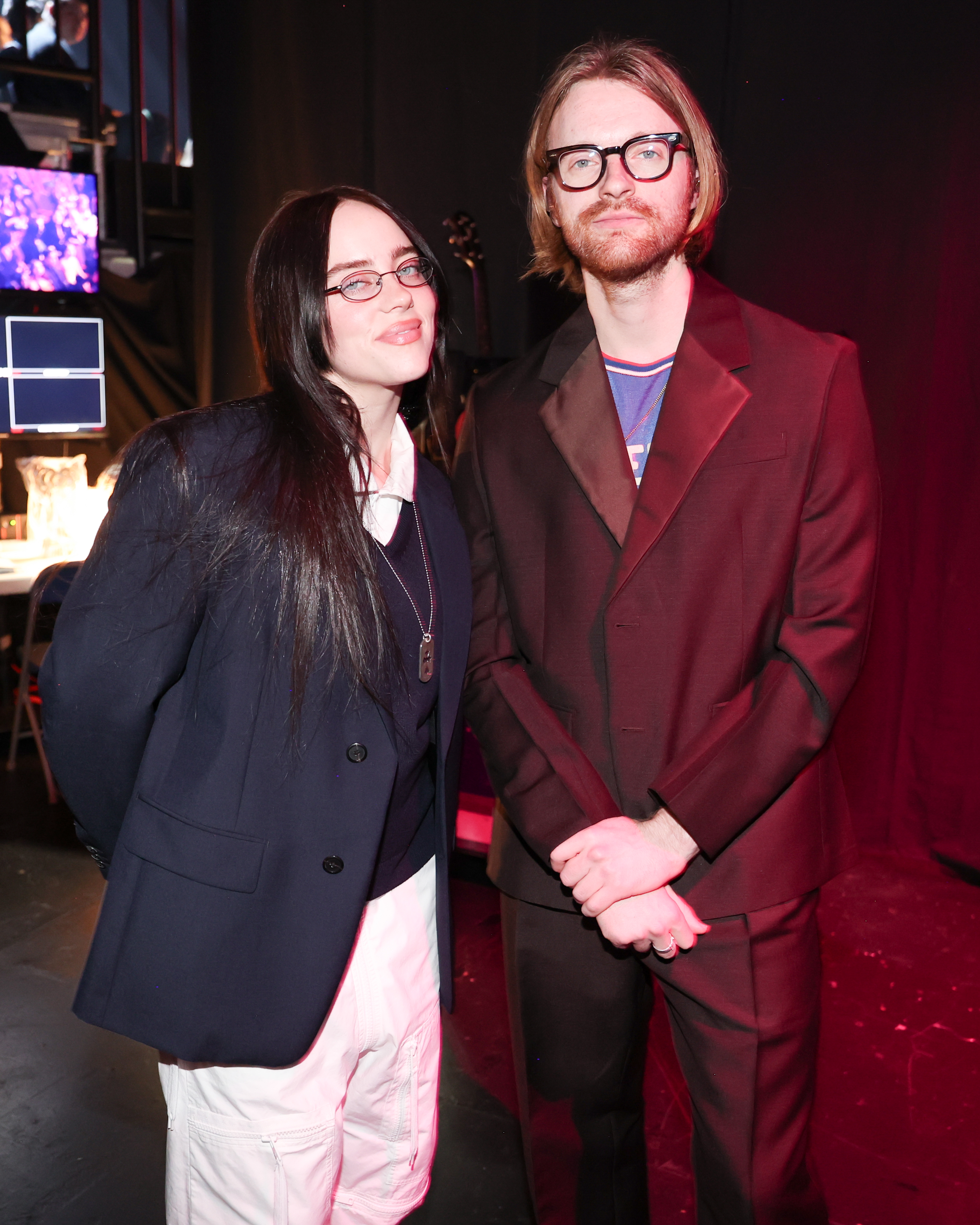 Two individuals pose together. The woman wears a blazer over a shirt and pants. The man wears a suit with glasses. Both smile slightly