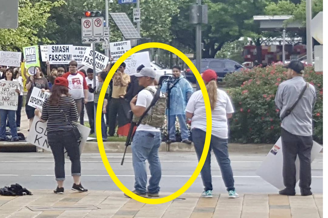 Protest scene with people holding signs. One individual in a vest and cap stands with a camera, capturing the event