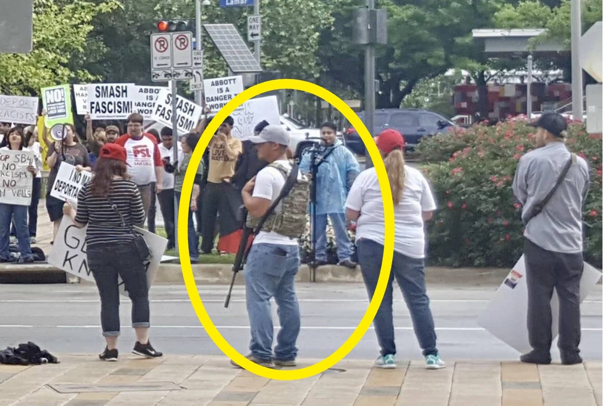 Protest scene with people holding signs. One individual in a vest and cap stands with a camera, capturing the event