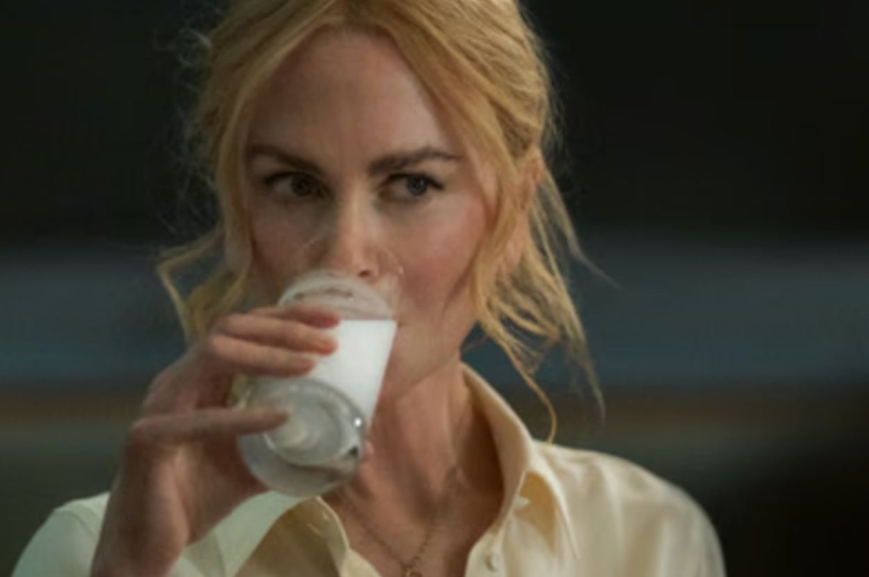 Person drinking from a glass of milk, wearing a light button-up shirt, looking contemplative