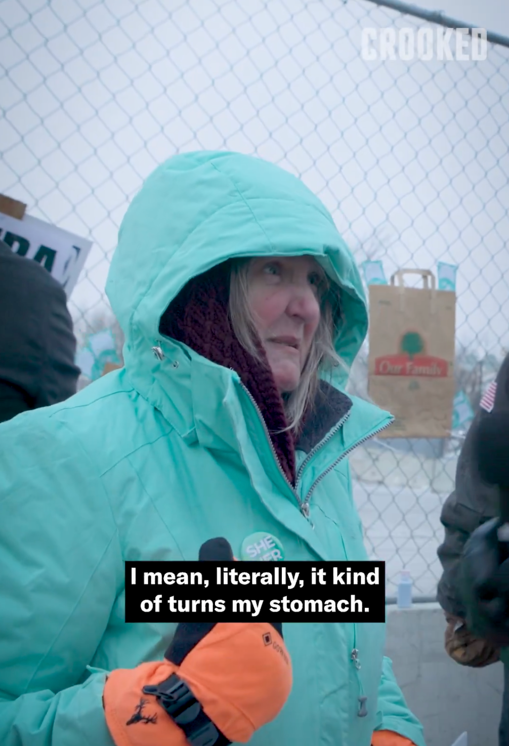 Person i blågrønn frakk og hette holder en mikrofon, snakker utenfor ved et gjerde, med teksten: 