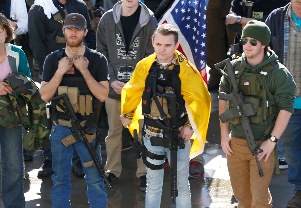 Tre personer står og holder rifler, med taktisk utstyr og et flagg synlig i bakgrunnen. De fremstår som seriøse og engasjerte i en hendelse