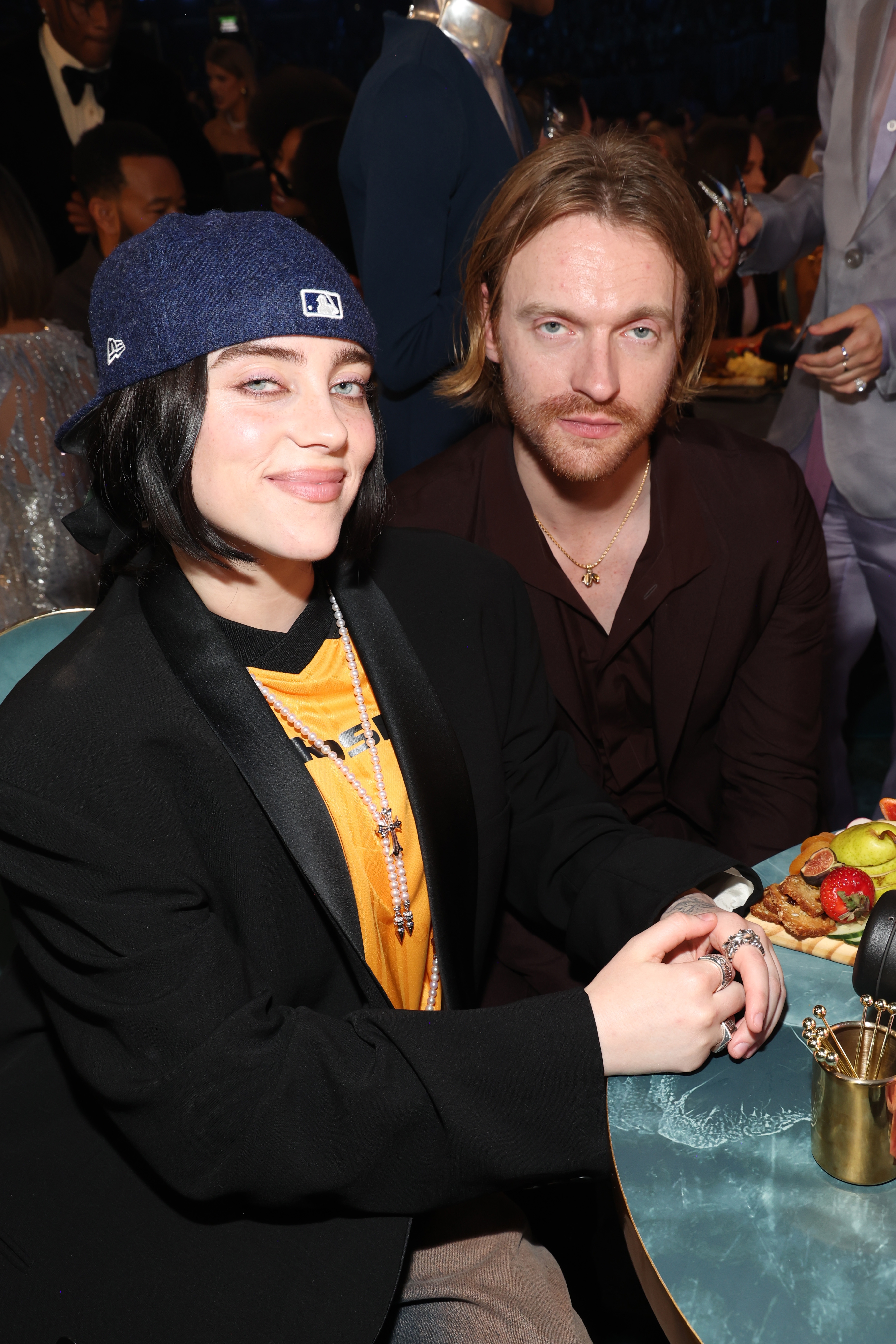 To personer som sitter på en formell begivenhet, den ene iført en uformell jakke og hatt, den andre i en mørk dress med et stjernekjede, smilende til kameraet