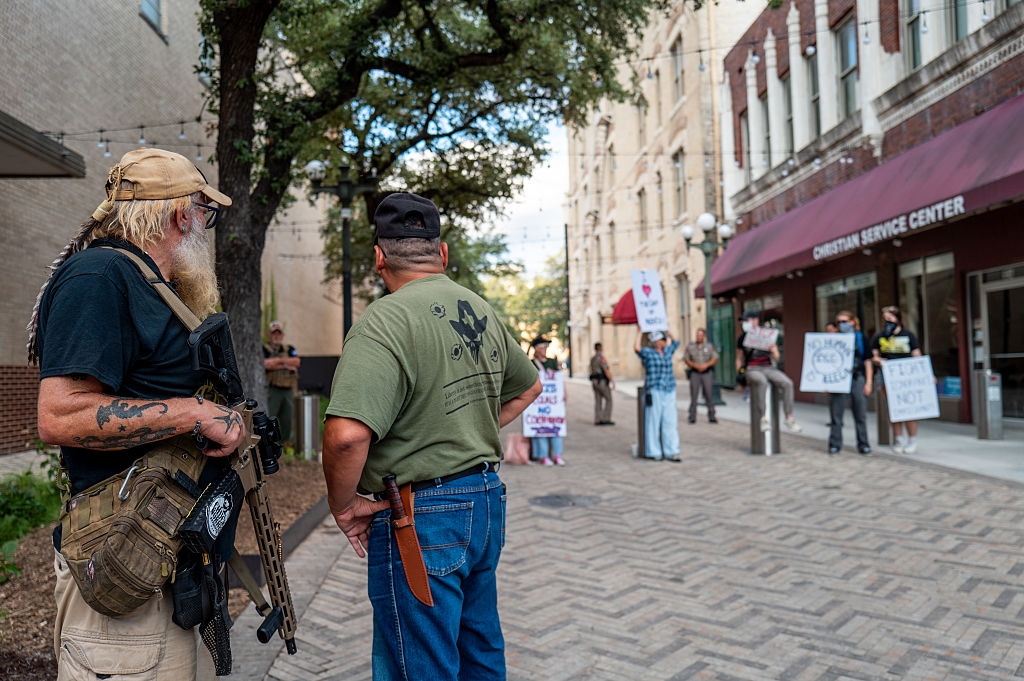 To menn med taktisk utstyr observerer en protest i urbane omgivelser, der folk holder skilt som uttrykker ulike budskap