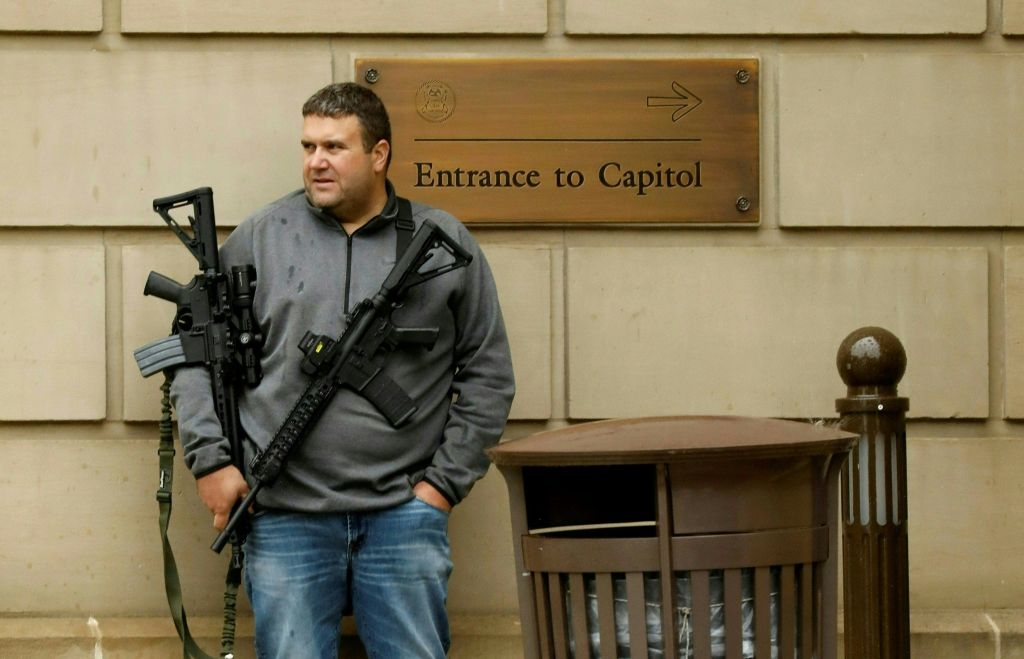 Mann som holder en rifle lener seg tilfeldig mot en vegg i nærheten av et skilt med teksten «Inngang til Capitol»; ved siden av en søppelbøtte