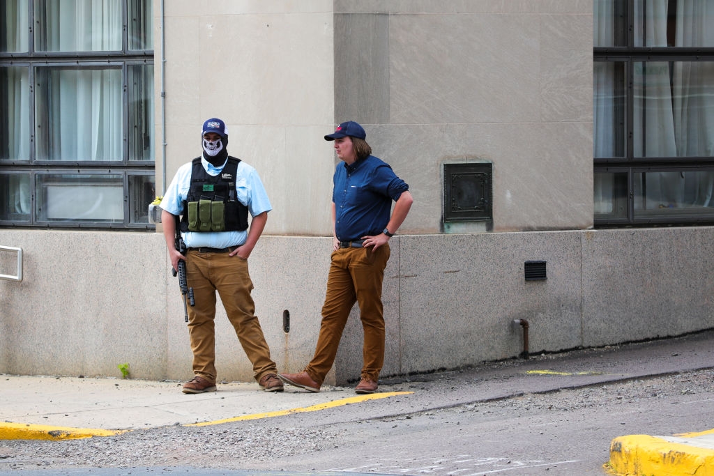 To menn iført uformelle klær og caps står på et fortau i byen. En mann bærer en taktisk vest og maske og holder en rifle; de ser ut til å være i samtale