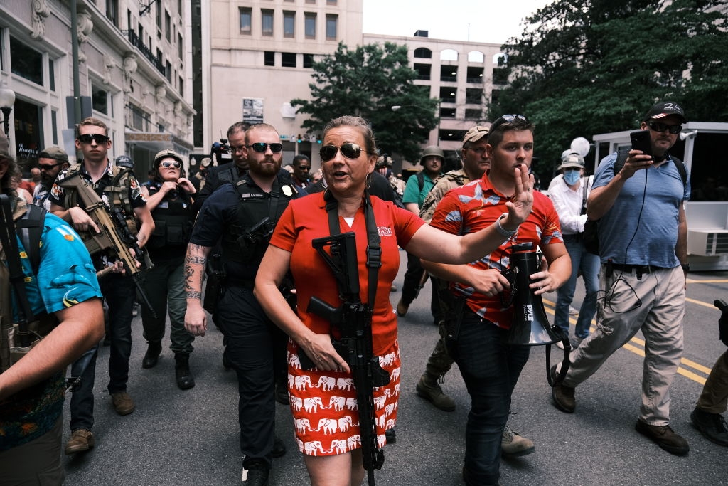 En kvinne leder en gruppe i en gateprotest, iført et mønstret skjørt og holder en rifle, mens andre rundt henne også holder skytevåpen