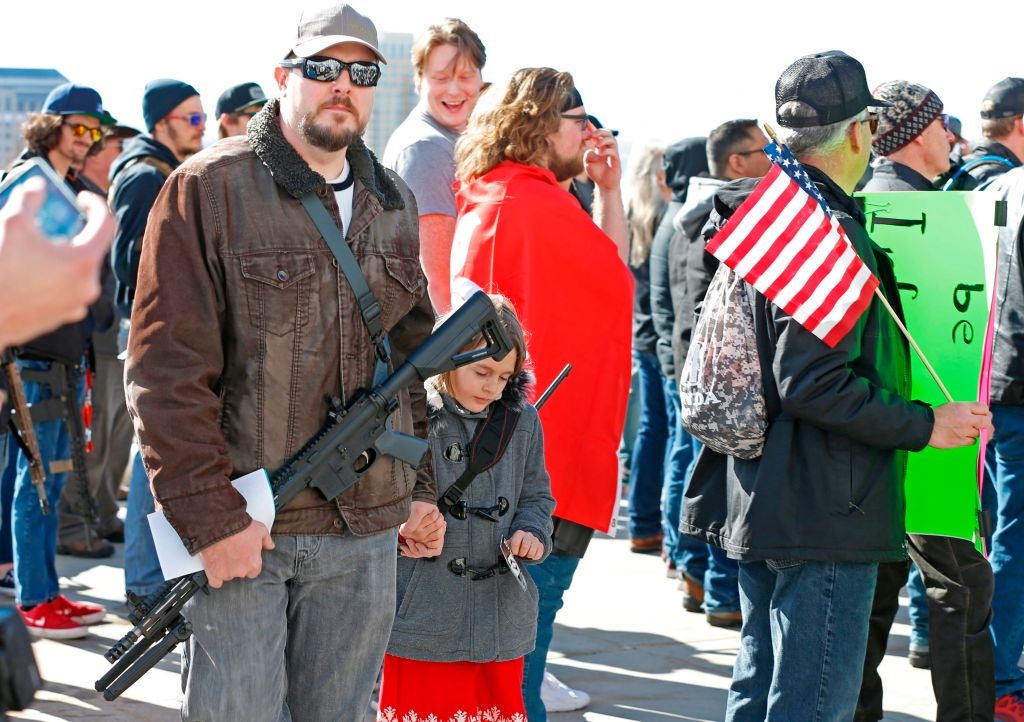 En mann og et barn i en folkemengde på et møte, begge holder rifler. Barnet har på seg en kjole, og en deltaker holder et amerikansk flagg