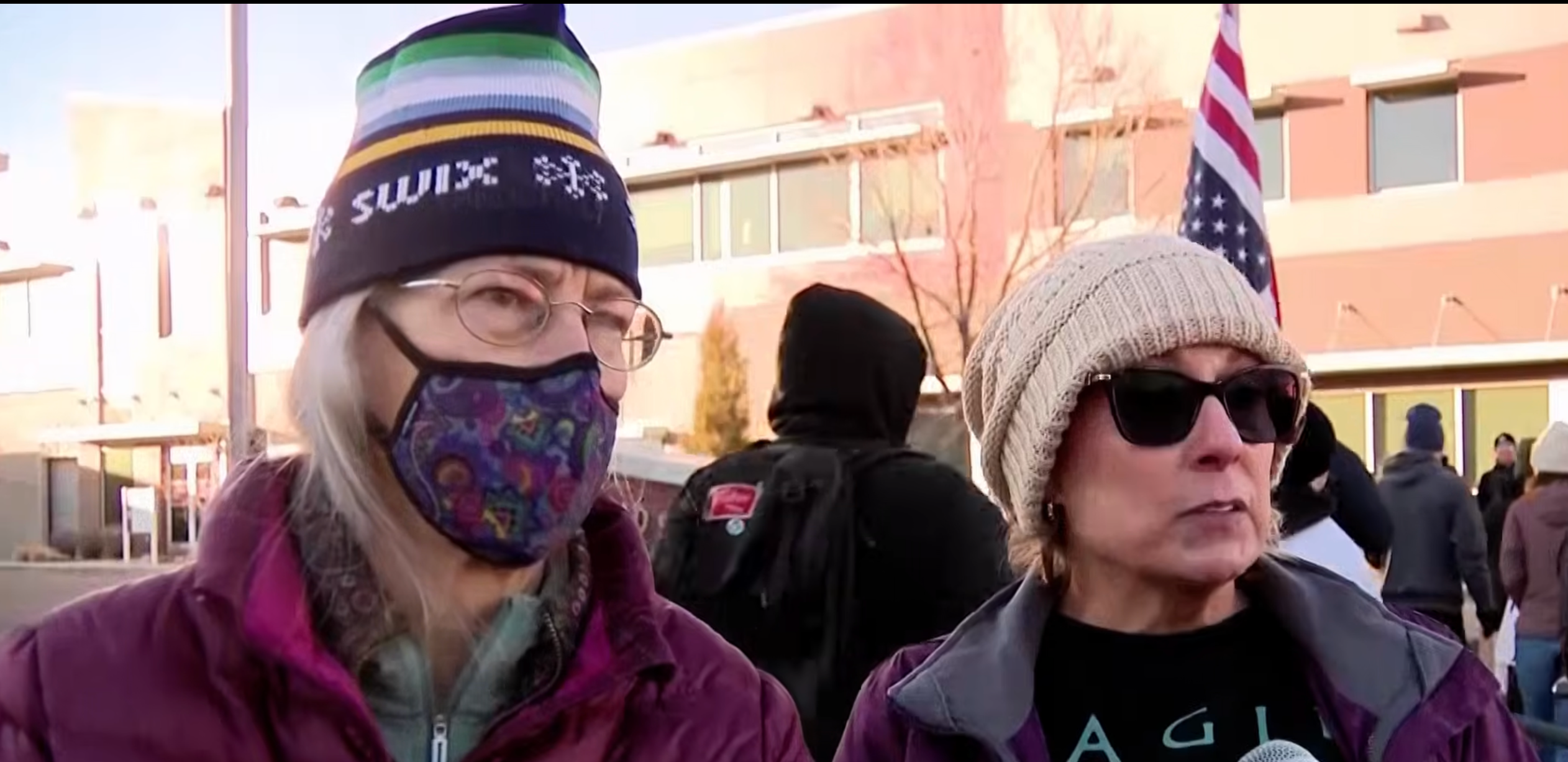 To kvinner med vinterhatter og frakker snakker utenfor. Et amerikansk flagg er synlig i bakgrunnen