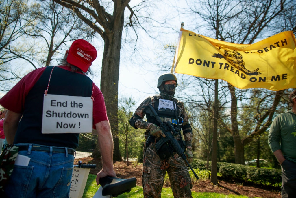 Demonstranter utendørs, en i taktisk antrekk som holder en rifle og «Don't Tread on Me" flagg; en annen har på seg en 