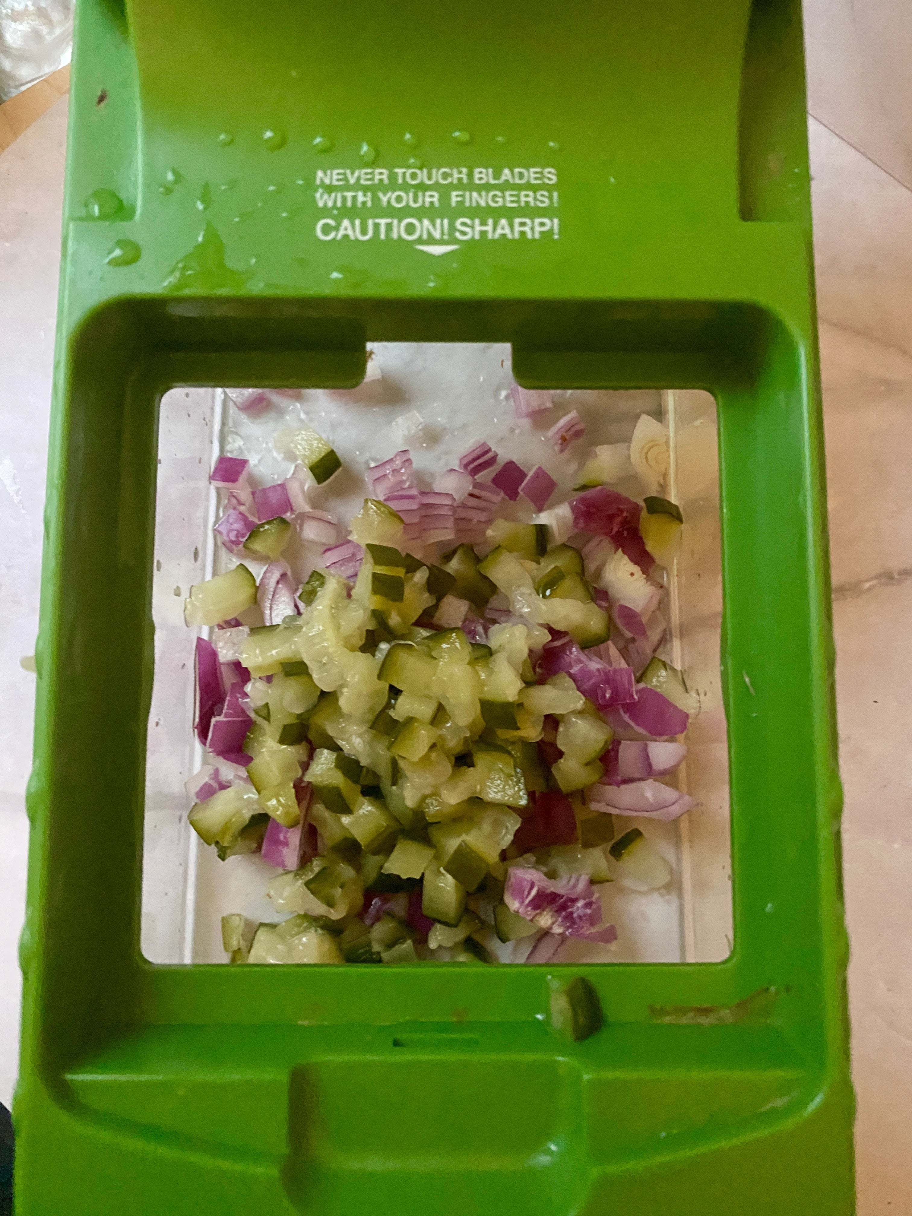 Chopped red onions and pickles are seen inside a green food chopper. Text warning: "Never touch blades with your fingers! Caution! Sharp!"