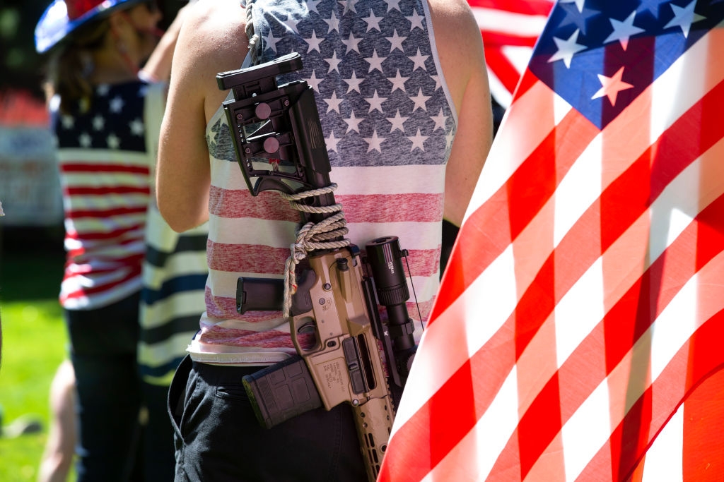 Person med rifle på ryggen, iført stjerne- og stripemønstrede klær nær det store amerikanske flagget