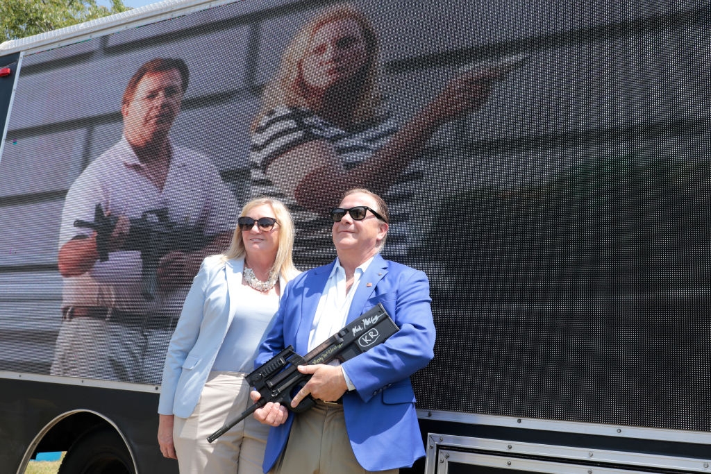 To personer står med en rifle og en pistol ved siden av et stort kjøretøy med bildet deres med de samme våpnene, begge med solbriller