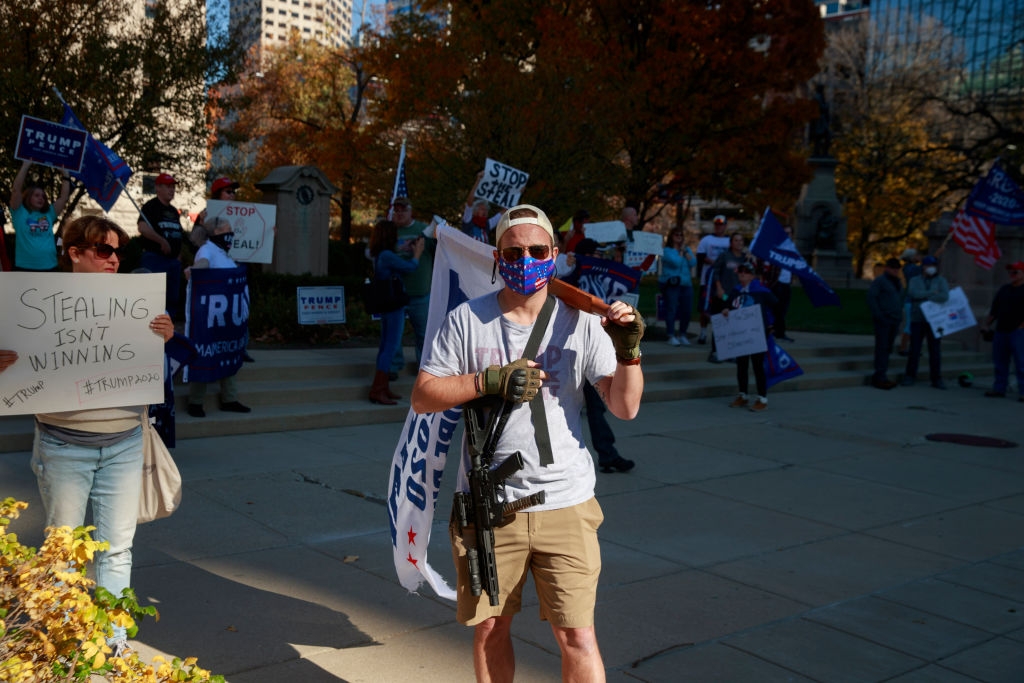 Demonstranter samlet seg utendørs, noen holdt skilt med politiske budskap. En person i t-skjorte og shorts bærer et stort flagg og bærer en ansiktsmaske