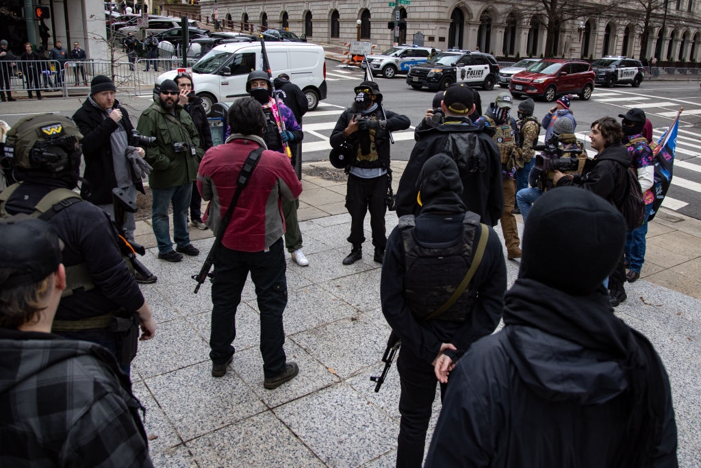 Gruppe individer, noen med taktisk utstyr, samlet seg på et gatehjørne i byen med politibiler i bakgrunnen