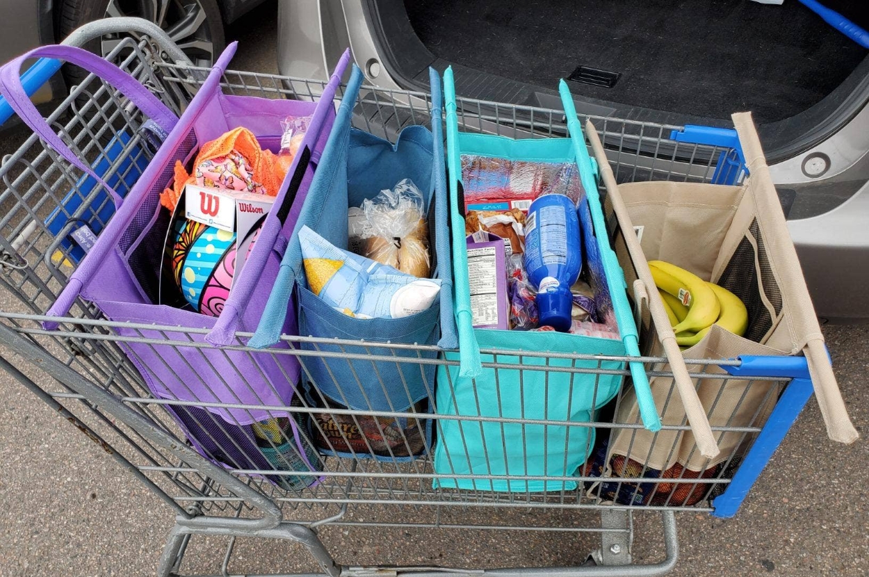 Shopping cart with reusable bags filled with groceries, including bananas, chips, and bottled items, beside an open car trunk