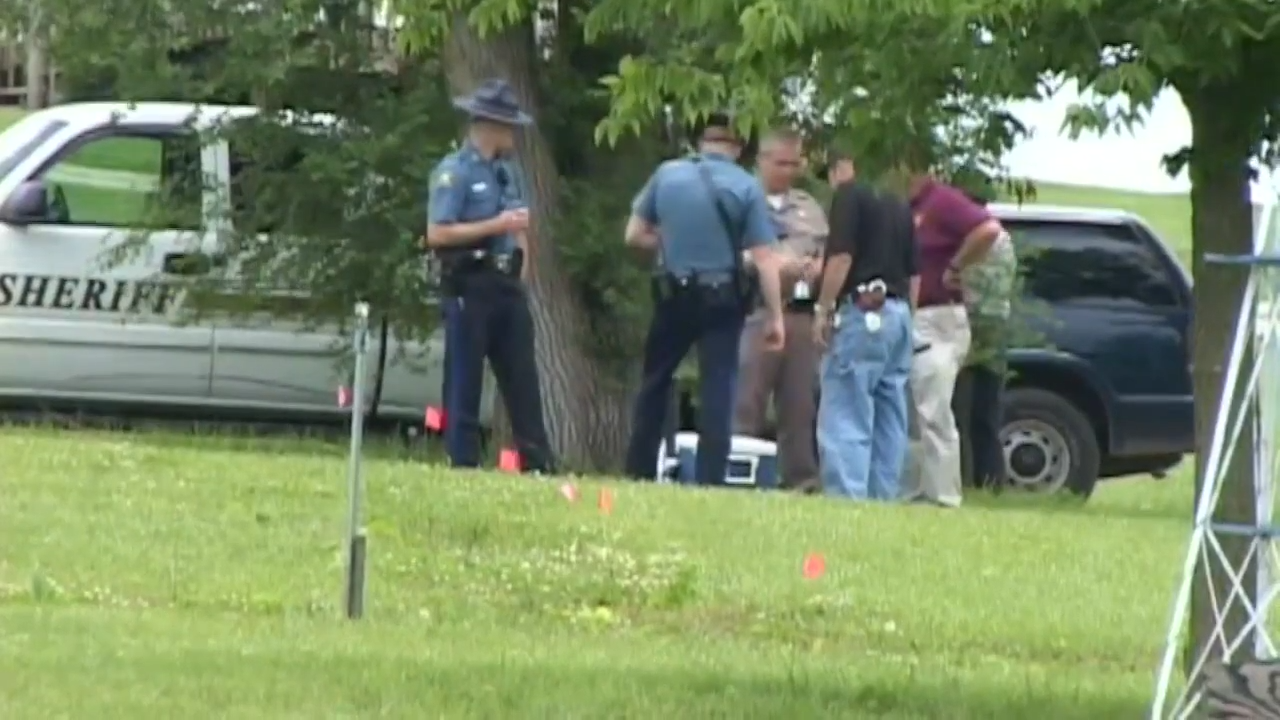 Politibetjenter undersøker en scene nær en lensmanns kjøretøy og trær i et gresskledd område