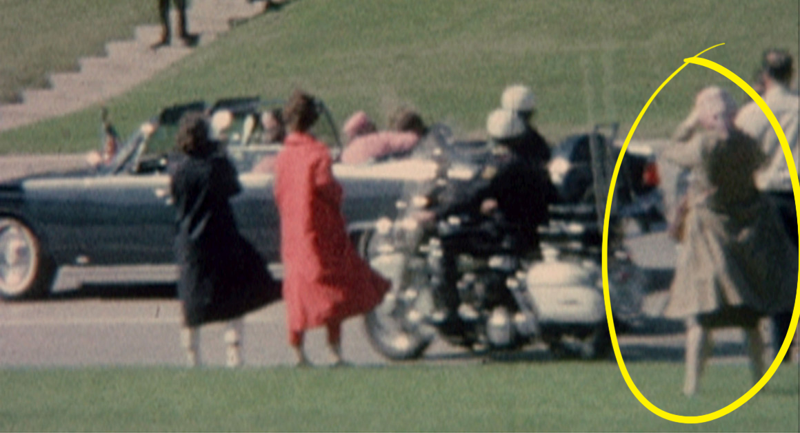 Folk står på gressletter nær kortegen. En person i frakk og hatt er omringet i gult