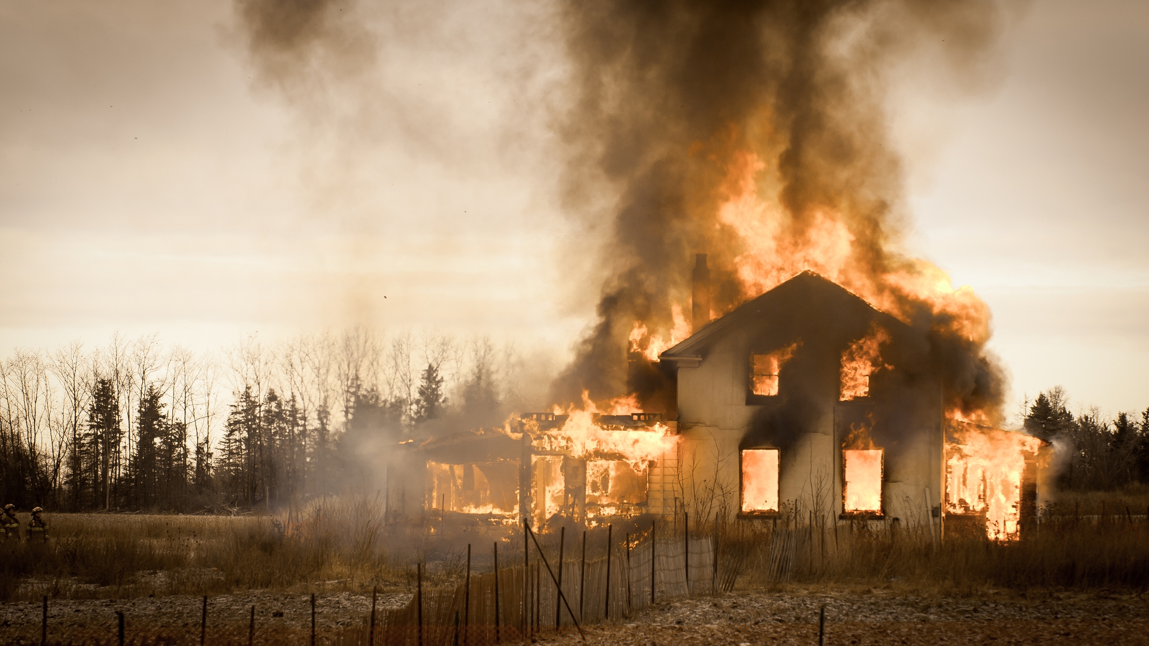 Et hus i et landlig område er oppslukt av flammer, med tykk røyk som stiger opp mot himmelen