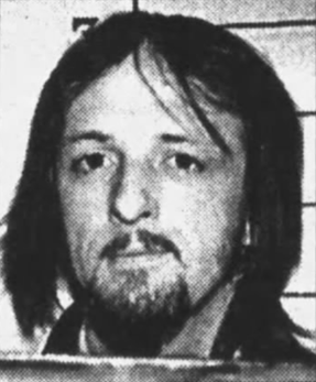 I don’t know who this person is, but a man with shoulder-length hair and a mustache looks ahead with a neutral expression in a black and white photo