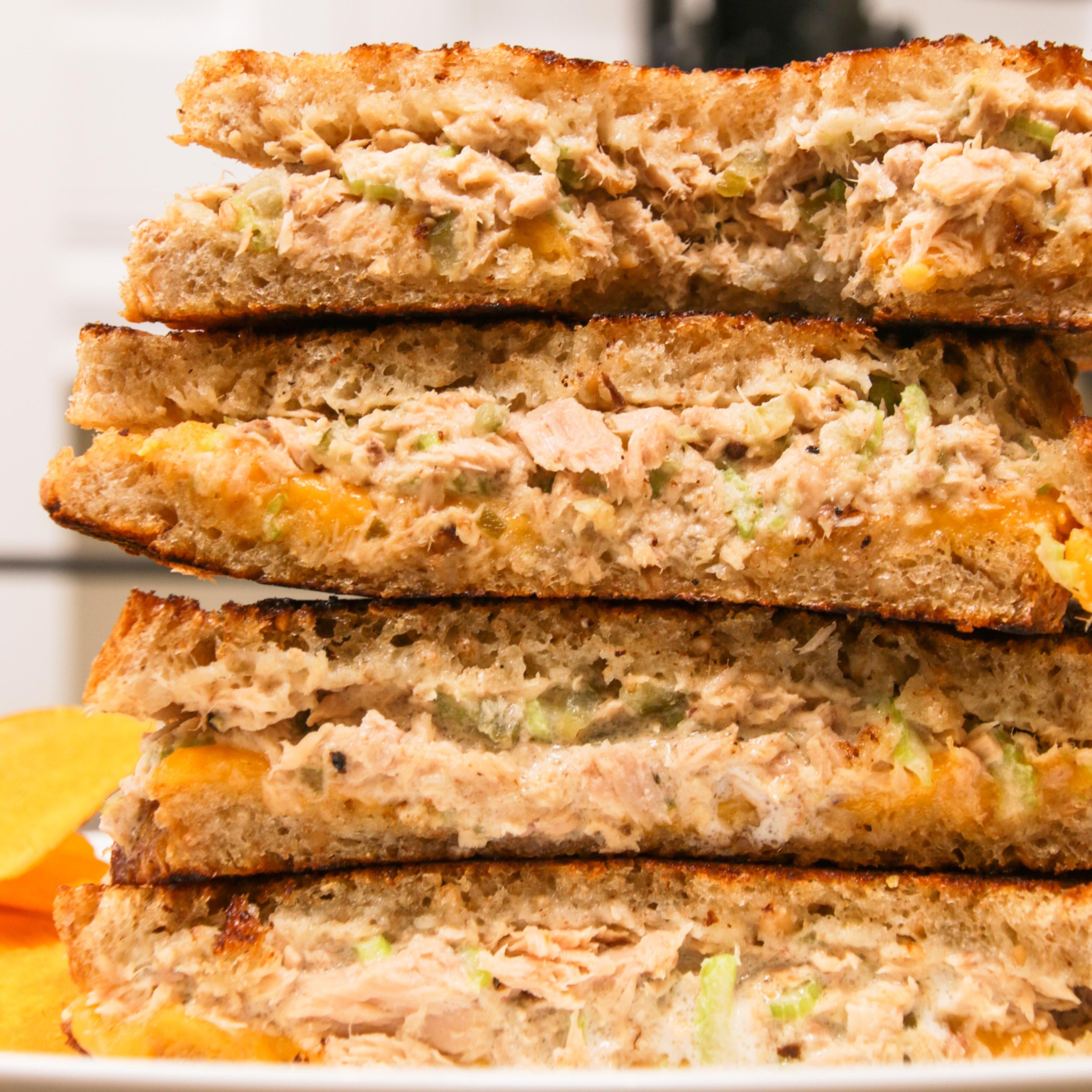 Stack of tuna melt sandwiches with grilled bread, melted cheese, and tuna salad filling, placed on a white plate with potato chips beside it