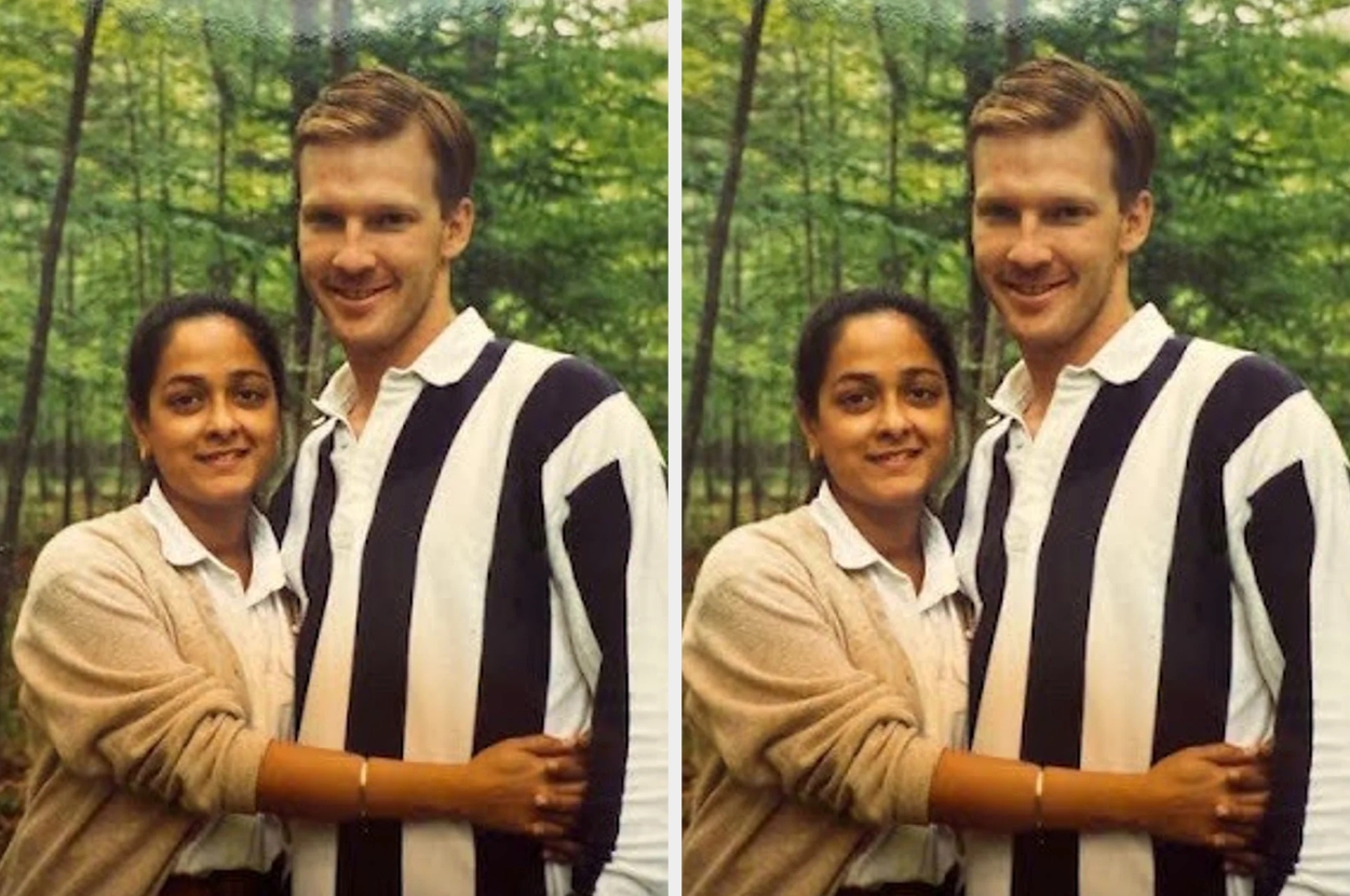 A couple stands closely in front of a forested background, smiling warmly at the camera