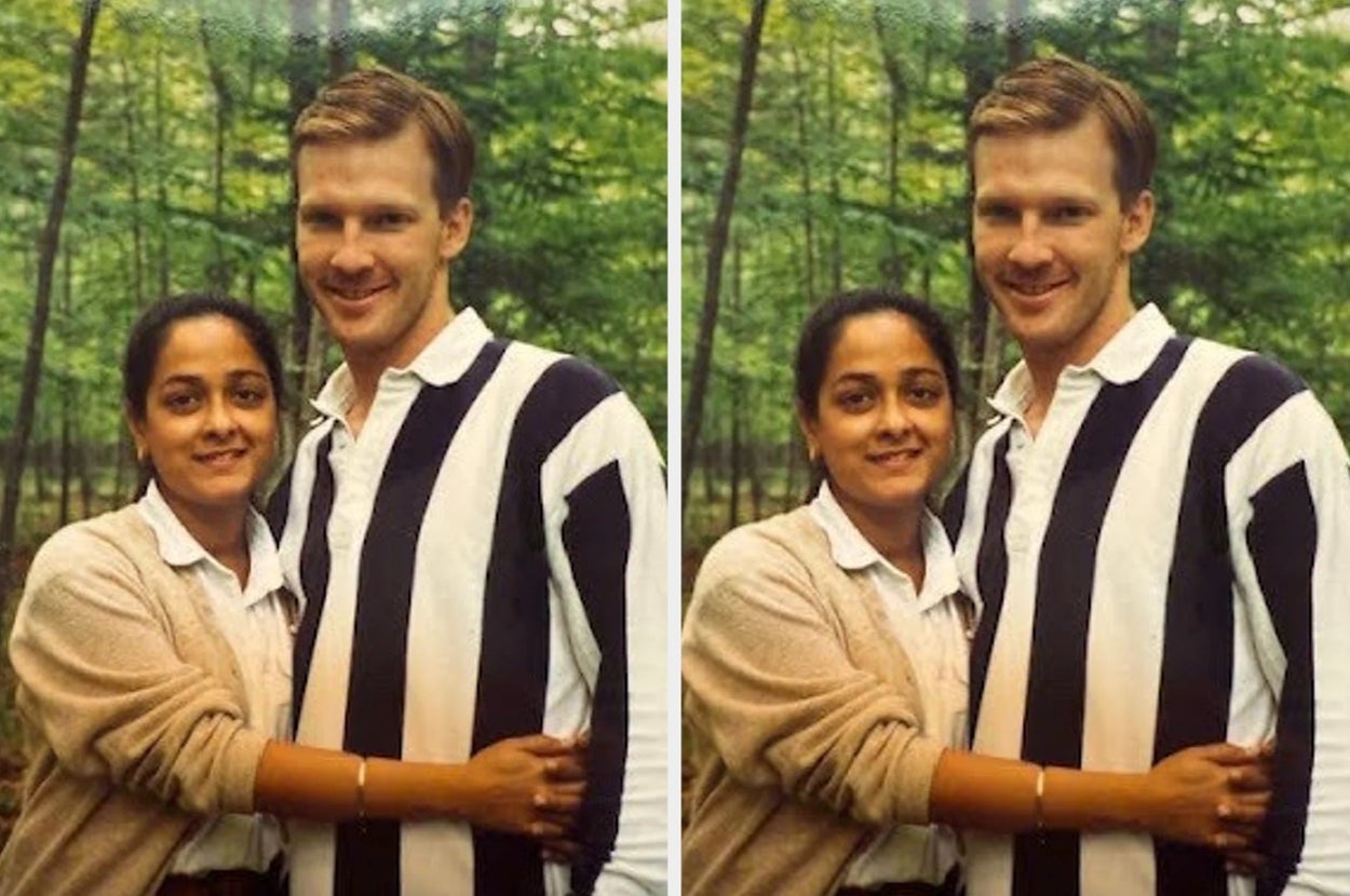 A couple stands closely in front of a forested background, smiling warmly at the camera
