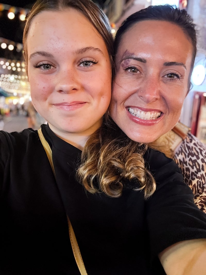 Two people smiling and posing for a selfie at an outdoor event. The atmosphere is cheerful, with decorative lights in the background