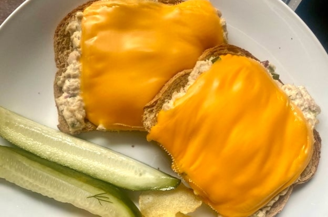 Two slices of toast with tuna salad, each topped with a slice of melted cheese, served with pickle spears on a plate