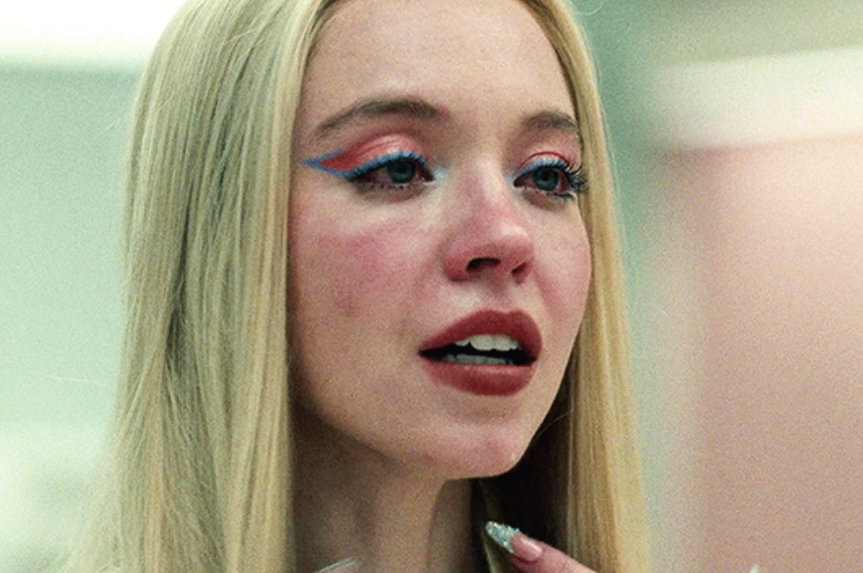 A woman with long blonde hair looks emotional, wearing bold eye makeup and long nails