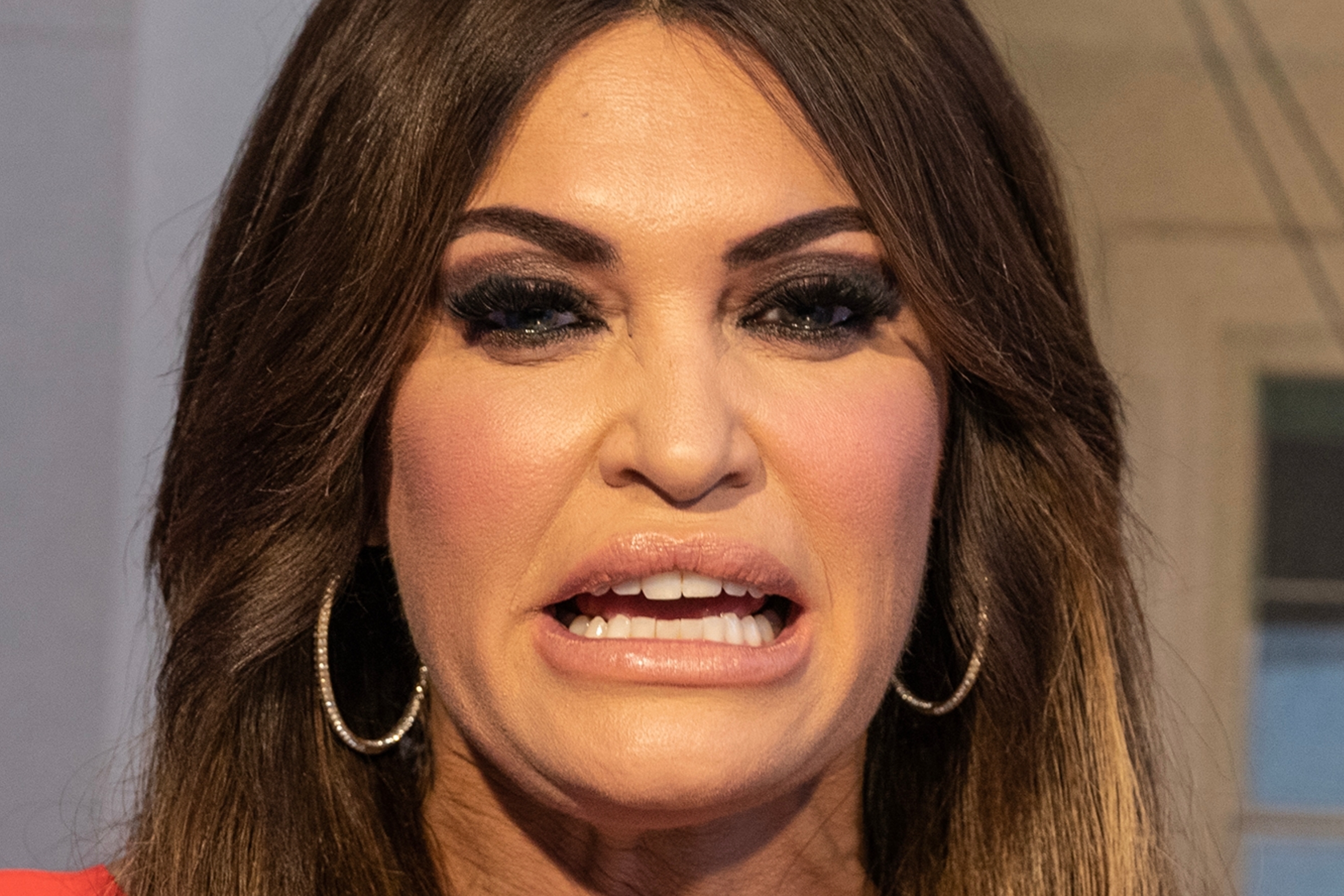 En person som gestikulerer med hendene hevet, iført en tilpasset kjole og ringøredobber, ser ut til å snakke eller uttrykke et poeng i en innendørs setting