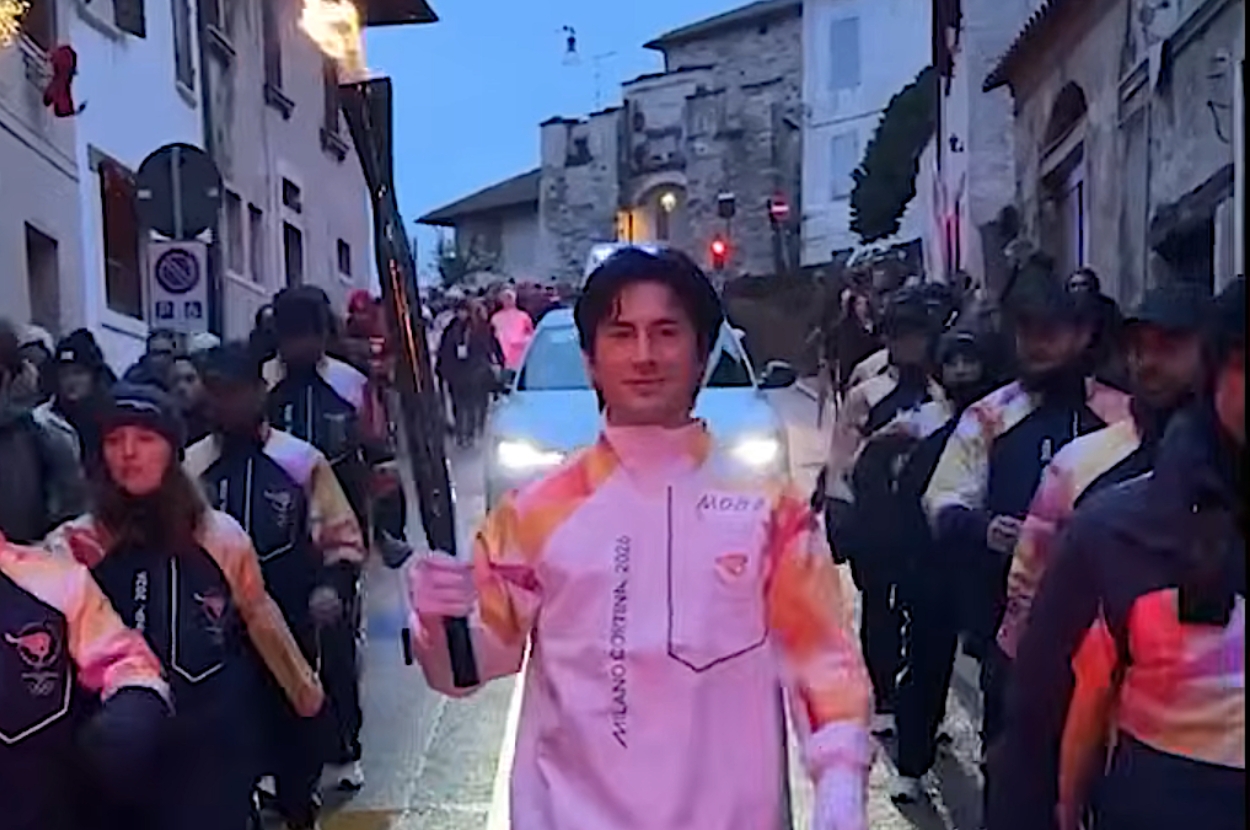 Hudson Williams holding a torch, leading a group during a parade along a street lined with spectators and vehicles behind him