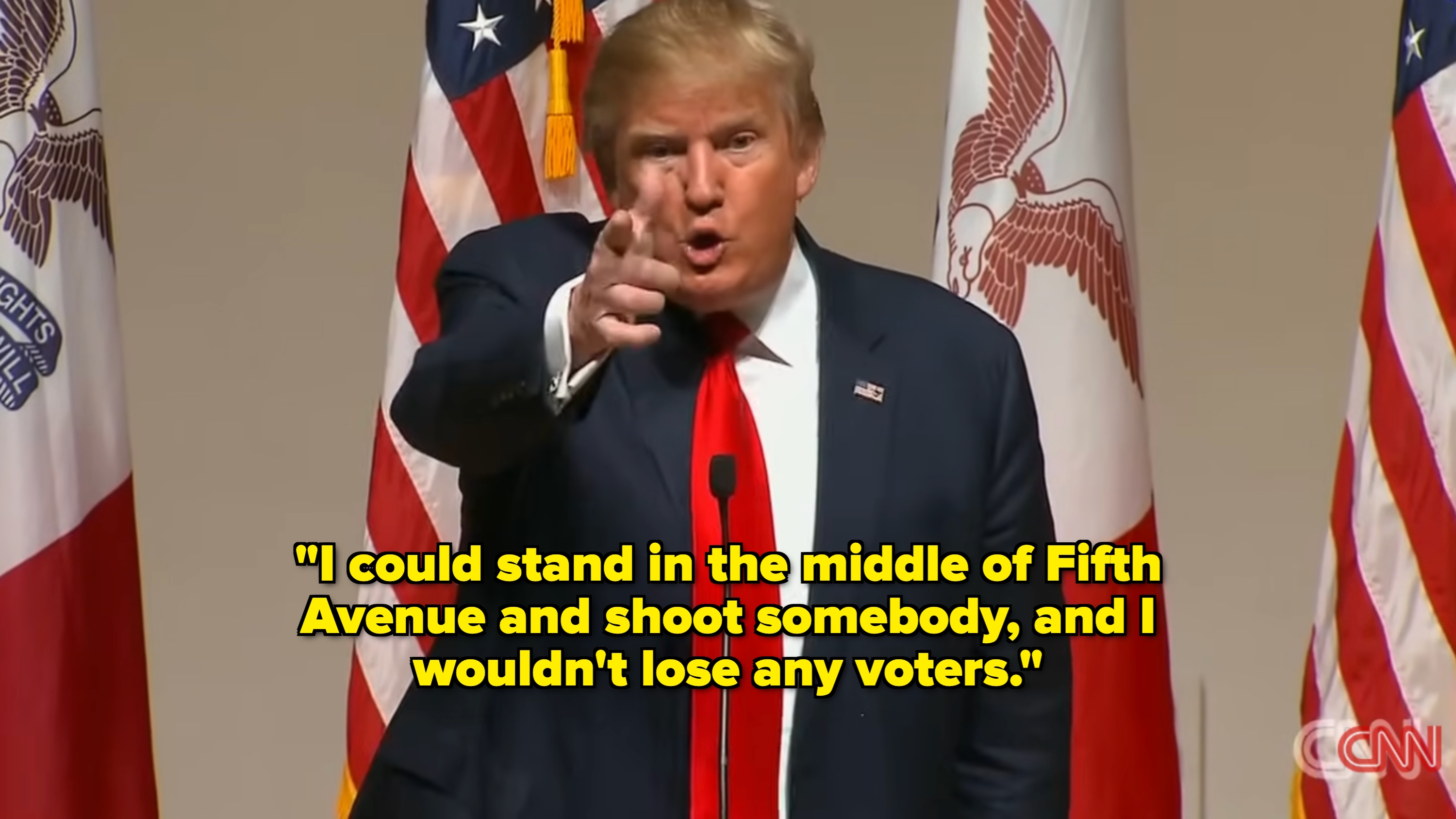 En person i dress peker fremover, snakker ved et podium, flankert av amerikanske og andre flagg. Innstillingen virker formell