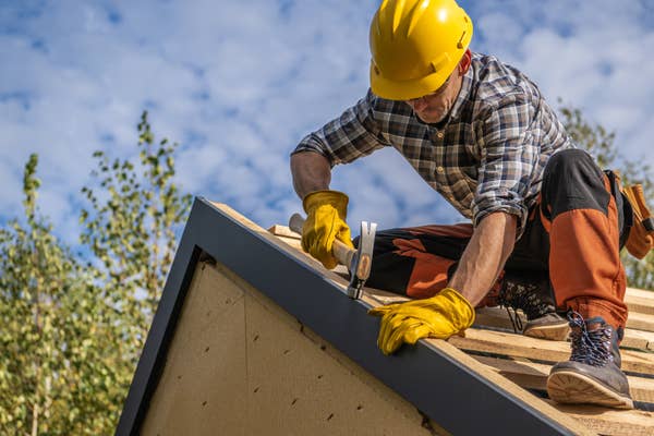 A construction worker in a helmet and gloves nails a panel on a roof, demonstrating careful craftsmanship and focus
