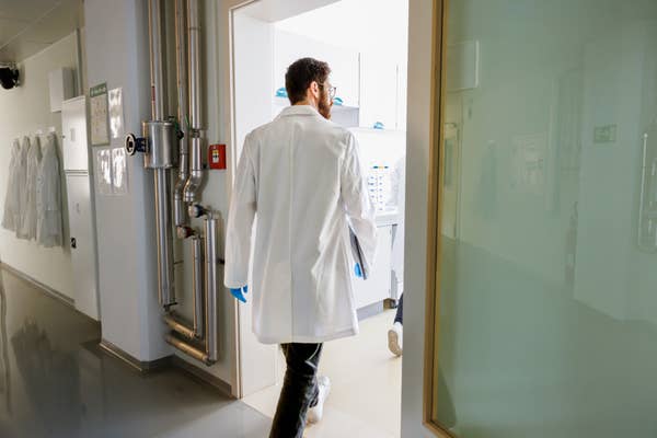Person in a lab coat enters a laboratory with stainless steel pipes visible, suggesting a professional science or research setting