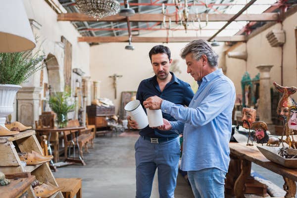 Two men in a large, rustic store discussing a ceramic item, one pointing while the other holds it, surrounded by various decorative objects