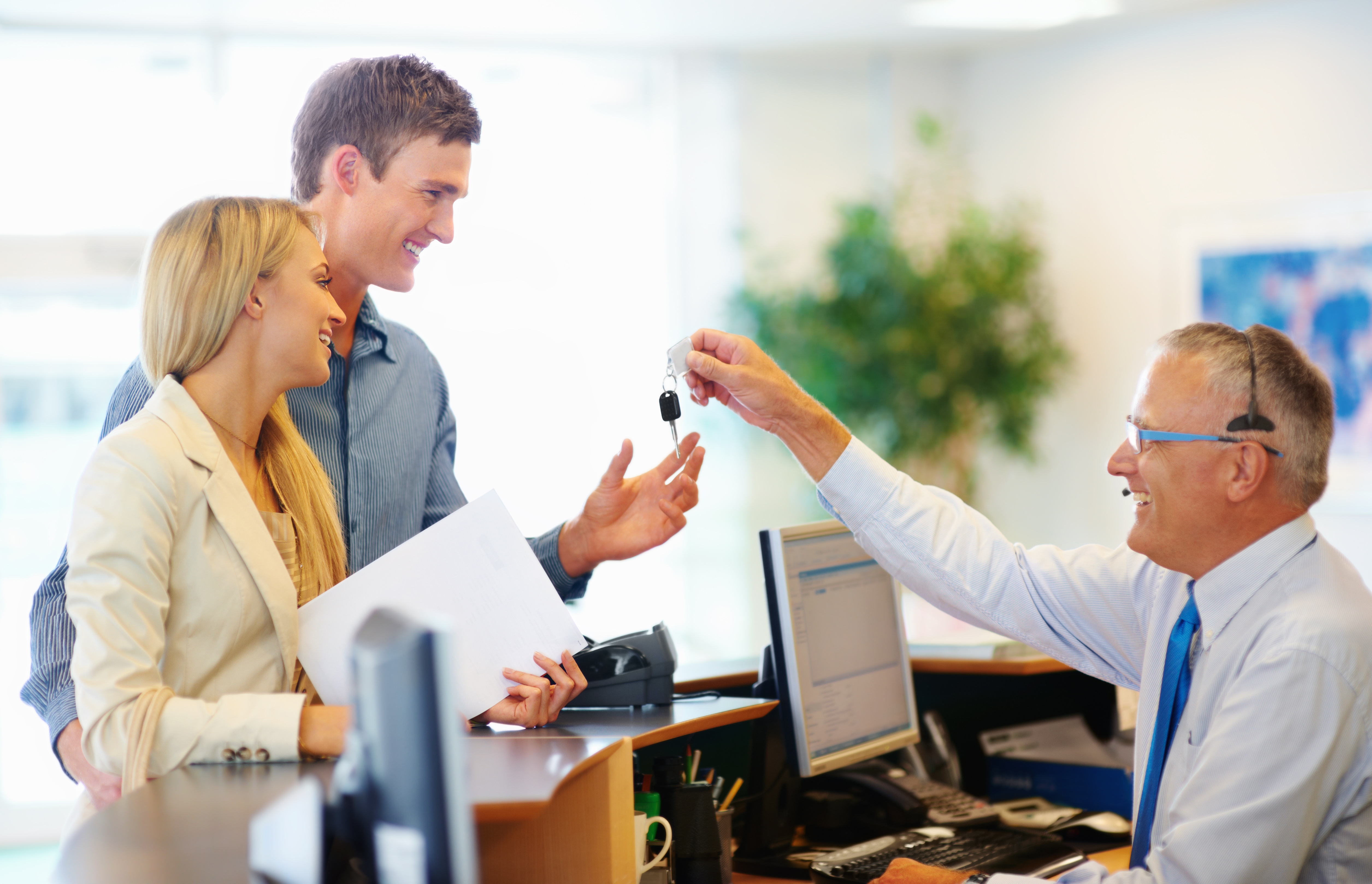 Smiling couple receives car keys from a dealership employee across a desk, symbolizing a new purchase or lease