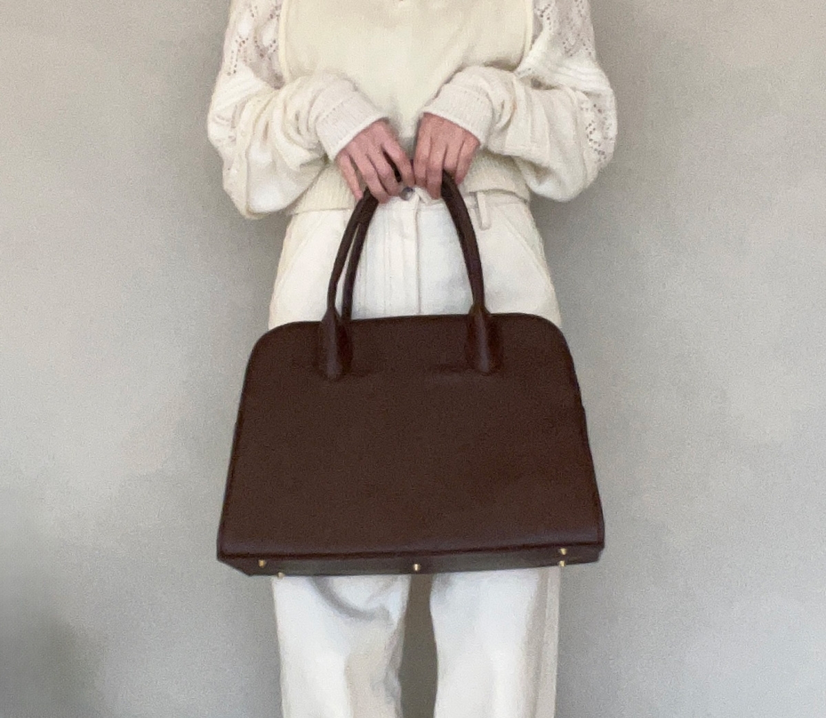 Person holding a large, dark handbag with a structured design, wearing a light sweater with textured sleeves and high-waisted pants