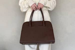 Person holding a large, dark handbag with a structured design, wearing a light sweater with textured sleeves and high-waisted pants