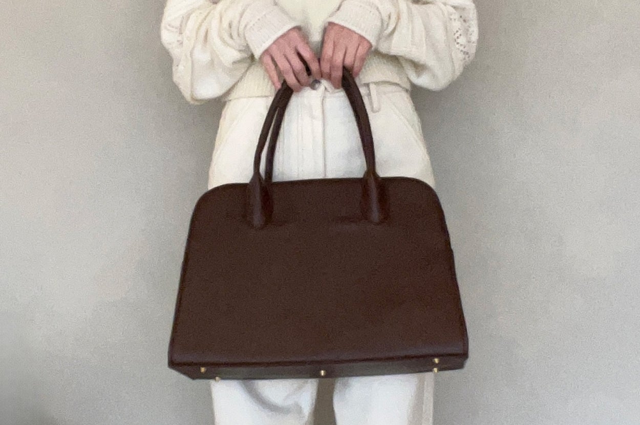 Person holding a large, dark handbag with a structured design, wearing a light sweater with textured sleeves and high-waisted pants