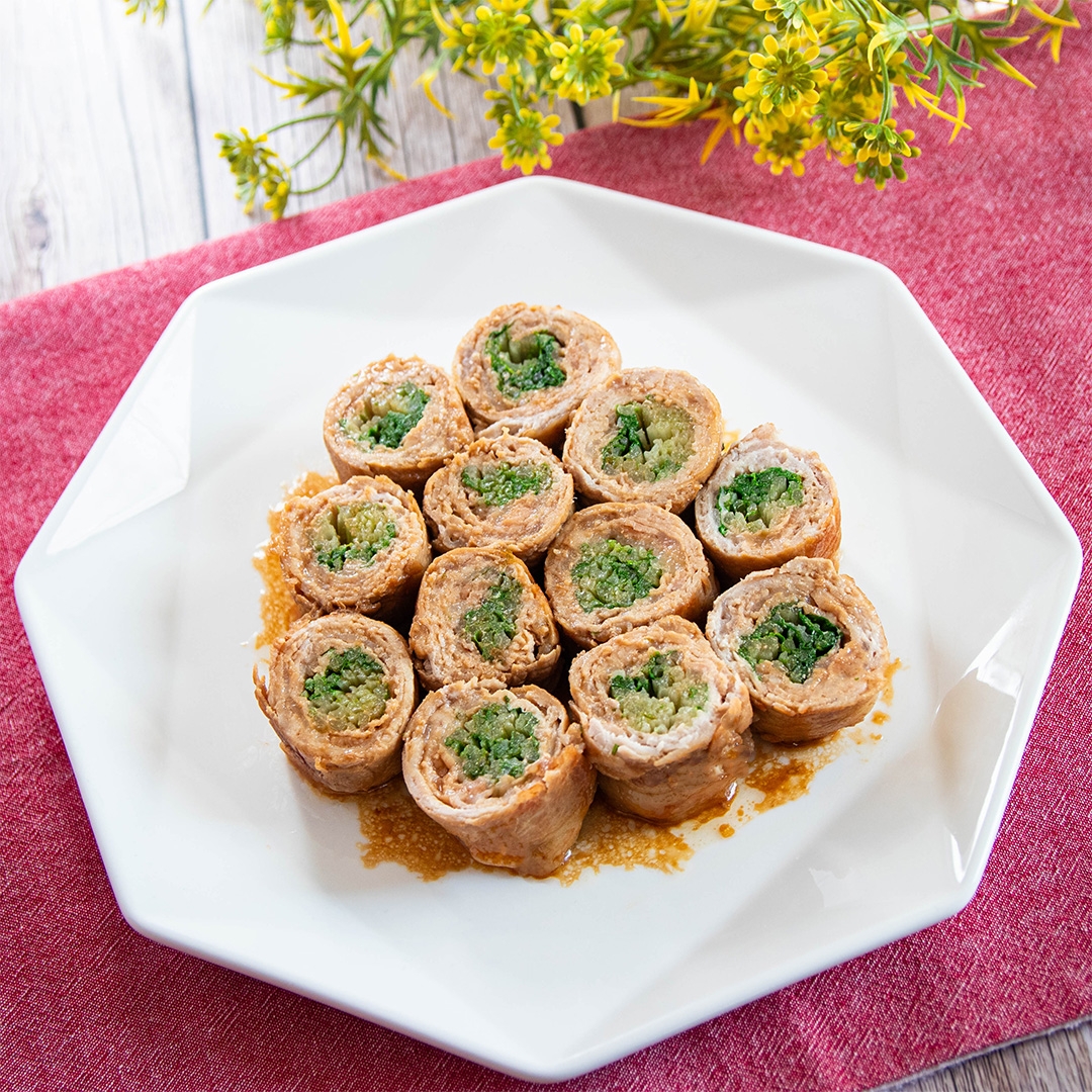 八角形の白い皿に盛り付けられた野菜と肉の巻き物料理。料理の下には赤いランチョンマット。花が飾られている。